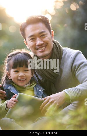 Außenportrait eines asiatischen Vaters und einer asiatischen Tochter, die auf Gras sitzen und lächelnd auf die Kamera blicken Stockfoto