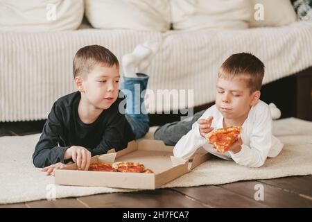 Zwei Jungen liegen auf dem Boden und essen Pepperoni Pizza aus einer Schachtel. Vertikaler Rahmen. Zwei Jungen liegen auf dem Boden und essen Pepperoni Pizza aus einer Schachtel. Stockfoto