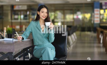Selbstbewusste und erfolgreiche junge Geschäftsfrau, die im Büro sitzt und eine Kaffeetasse hält. Stockfoto