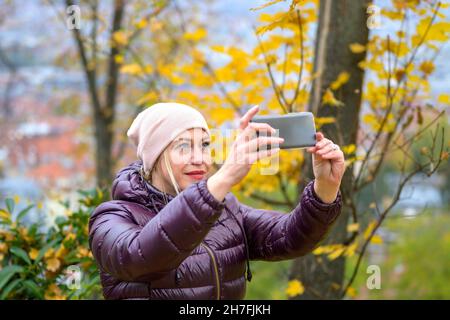 Frau mittleren Alters in einer warmen violetten Jacke und einem rosa Hut, die in der Herbstsaison ein Foto der wunderschönen Natur in einem Park macht und ihr Mobiltelefon in beiden Händen hält Stockfoto