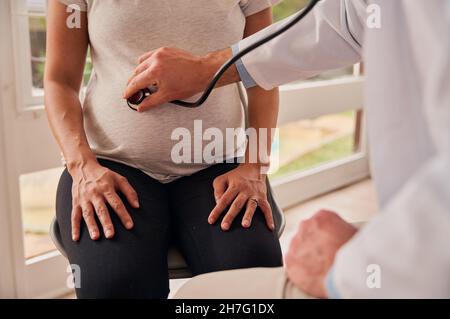 Kaukasische Schwangere, die in der Klinik sitzt, während ein männlicher Arzt den Schwangeren mit Stethoskop untersucht Stockfoto