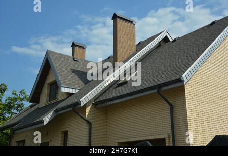 Eine Nahaufnahme eines Doppelkomplexes, das Dach eines Backsteinhauses, das mit grauen Asphaltschindeln, Faszien und Soffit bedeckt ist, mit einem Dachfenster Stockfoto