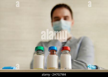 Der maskierte Mann sieht sich die Flaschen verschiedener Impfstoffe an, das leere weiße Etikett auf der Flasche für Text und Design. Die Wahl des Impfstoffs. Stockfoto