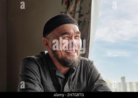 Porträt eines glücklichen älteren asiatischen Mannes mit Bart und Totenkopf am Fenster sitzend und lächelnd. Stockfoto
