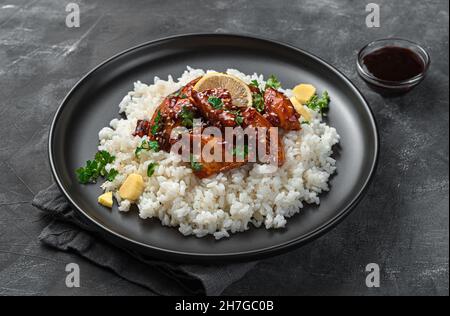 Eine Portion Chicken Teriyaki mit Reis, Sesam und frischer Petersilie auf dunklem Grund. Stockfoto