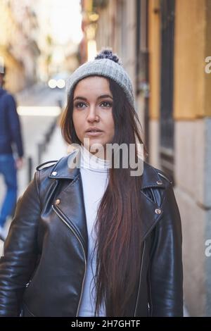 Junge Frau mit langen dunklen Haaren, die Lederjacke und warmen Hut trägt und auf unscharfen Hintergrund auf der Straße der Stadt schaut Stockfoto