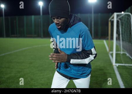 Bärtiger afrikanischer Fußballspieler in professioneller Sportuniform läuft während des Trainings entlang des Fußballfeldes Stockfoto