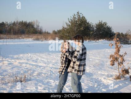 Ein fröhliches, verliebtes Teenager-Mädchen und ein etwa 16-17-jähriger Kerl in karierten Hemden und Jeans stehen im Schnee, umarmen sich und lachen fröhlich. Die Conce Stockfoto