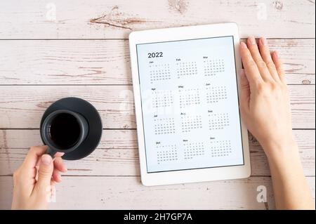 Tablet-Computer mit einer offenen App von Kalender für 2022 Jahre und Tasse Tee oder Kaffee in Frauenhand auf Holzhintergrund. Concept Business oder zu tun li Stockfoto