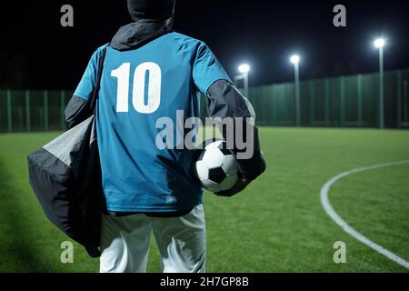 Rückansicht des Fußballspielers mit Tasche und Fußball, der sich in Richtung Grünfeld bewegt, während er trainiert Stockfoto
