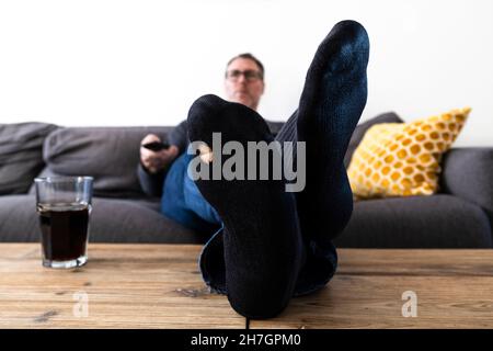 Erwachsener Mann mit einem Loch in seiner Socke, der Fernsehen schaute und die Fernbedienung benutzte Stockfoto