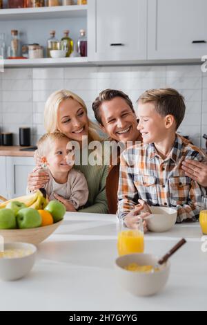 Fröhliche Eltern, die Kinder umarmen, während sie morgens in der Küche frühstücken Stockfoto