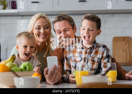 Fröhlicher Mann, der mit seiner Frau und seinen Söhnen ein Selfie auf dem Handy beim Frühstück in der Küche gemacht hat Stockfoto