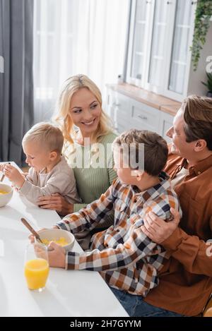 Fröhlicher Mann und Frau, die sich in der Nähe von Söhnen beim Frühstück mit Cornflakes angucken Stockfoto