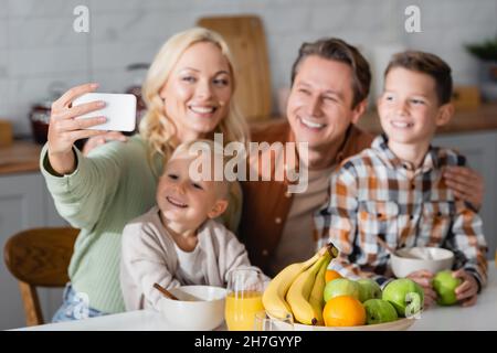 Fröhliche Frau, die während des Frühstücks Selfie mit der Familie in der Nähe von frischen Früchten am Handy gemacht hat Stockfoto