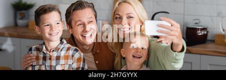 Fröhliche Frau, die Selfie auf dem Smartphone mit Ehemann und Söhnen in der Küche macht, Banner Stockfoto
