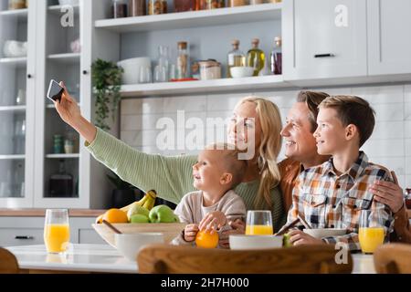 Glückliche Frau, die Selfie auf dem Handy macht, während sie mit der Familie frühstückt Stockfoto