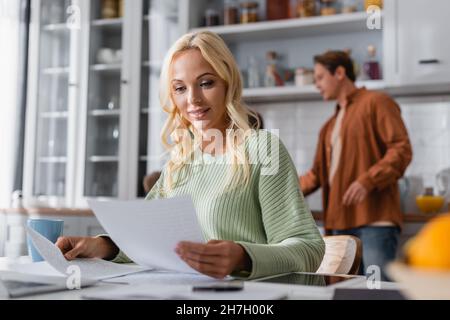 Frau, die sich das Dokument ansieht, während sie in der Küche in der Nähe einer verschwommenen Familie arbeitet Stockfoto