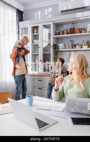 Mann, der mit Kindern in der Küche neben seiner Frau spielt, die in der Nähe des Laptops auf dem verschwommenen Vordergrund arbeitet Stockfoto