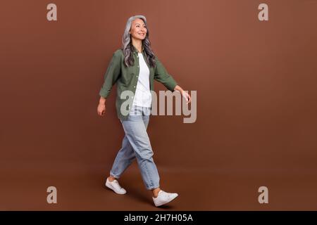 In voller Länge Foto von schönen älteren weißen Frisur Dame gehen tragen Khaki Hemd Jeans Sneakers isoliert auf braunen Hintergrund Stockfoto