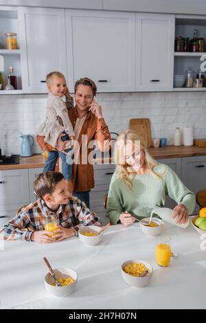 Frau gießt Milch in Cornflakes und Ehemann spricht während des Frühstücks mit Kindern auf dem Smartphone Stockfoto