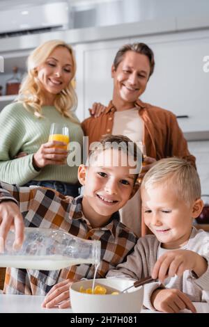 Lächelndes Kind, das die Kamera anschaut, während es Milch mit leckeren Cornflakes in die Schüssel gießt Stockfoto