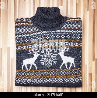 Blauer gestrickter weihnachtlicher Rollkragenpullover mit geometrischem Elch- oder Elchornament (auch Ugly Sweater genannt), gefaltet auf Holzhintergrund gelegt Stockfoto