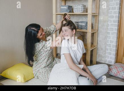 Frohe, liebevolle Familie. Eine Mutter kämmt die Haare ihrer Tochter, während sie in einem Zimmer auf dem Boden sitzt Stockfoto