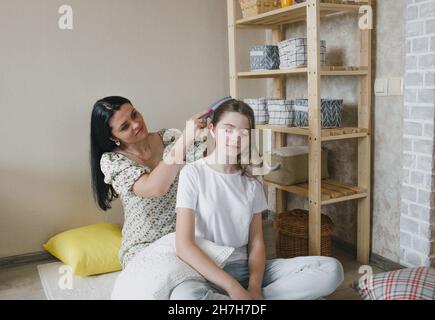 Frohe, liebevolle Familie. Eine Mutter kämmt die Haare ihrer Tochter, während sie in einem Zimmer auf dem Boden sitzt Stockfoto