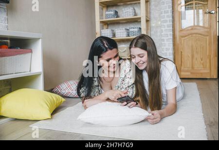 Eine fröhliche Mutter schaut sich ein Video auf ihrem Telefon an, während ihre lachende Tochter auf weichen Kissen auf dem warmen Boden des Hauses zusammenliegt. Stockfoto