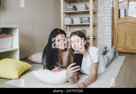 Eine fröhliche Mutter schaut sich ein Video auf ihrem Telefon an, während ihre lachende Tochter auf weichen Kissen auf dem warmen Boden des Hauses zusammenliegt. Stockfoto