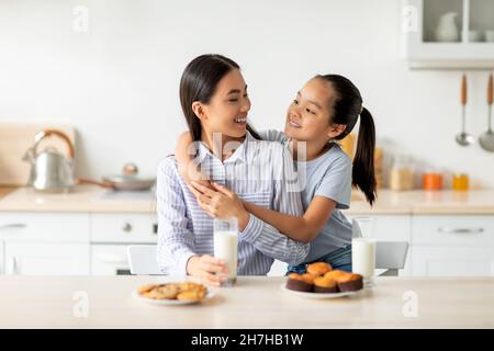 Nette asiatische Tochter umarmt ihre junge glücklich Mutter, während genießen hausgemachte Kekse und Kekse in der Küche Stockfoto