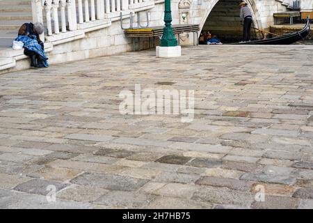 Eine obdachlose Frau auf einem Platz in Venedig. Zwei Touristen in einer Gondel fahren vorbei. Stockfoto