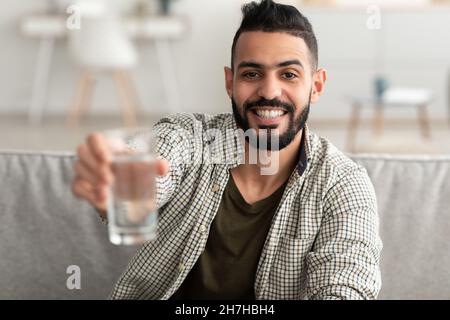 Hübscher junger Araber, der ein Glas frisches klares Wasser in der Hand hält und zu Hause lächelt Stockfoto