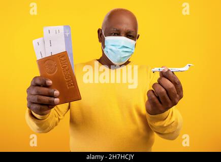 Älterer schwarzer männlicher Tourist in Gesichtsmaske mit Pass, Tickets und Spielzeugflugzeug, Blick auf die Kamera auf orangefarbenem Hintergrund Stockfoto