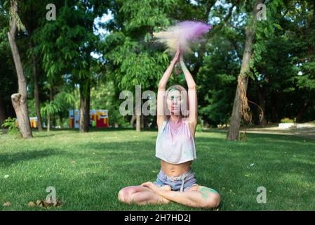 Eine blonde Frau meditiert im Park beim Holi-Festival, indem sie ihre Hände hochhebt und mehrfarbige trockene Farben streut. Meditation im Freien. Farbiger Puder Stockfoto