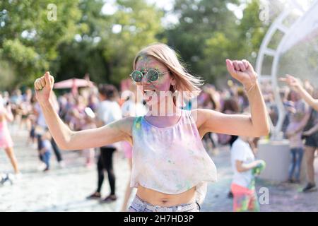 Verrückte kaukasische Mädchen auf Holi Fest Festival im Stadtpark im Sommer. Ein Farbenfest aus trockenen Farben und eine Blondine, die zur Musik tanzt. Geburtstagsfeier Konzept Stockfoto