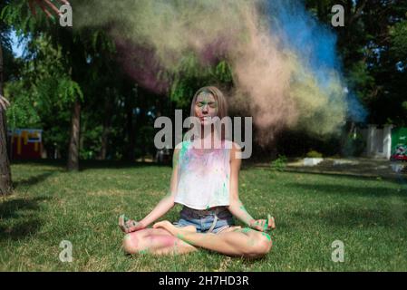 Junge Frau in Lotusstellung beim Holi-Festival meditiert in der Natur. Color Fest und Outdoor-Meditation. Mehrfarbiger Puder färbt das Gesicht. Stockfoto