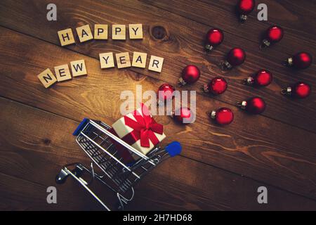 Geschenk in einem kleinen Karren, Beschriftung in Holzbuchstaben Frohes Neues Jahr und mehrfarbige Weihnachtskugeln auf einem hölzernen Hintergrund. Dunkler Weihnachtshintergrund Stockfoto