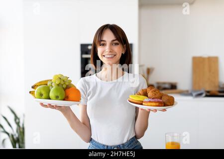 Gesund vs. Ungesund. Glückliche Frau hält Teller mit Obst und Desserts und wählt zwischen Gemüse und Süßigkeiten Stockfoto