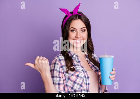 Foto-Porträt des lächelnden Mädchen zeigt Daumen hinter dem Rücken hält blauen Kunststoff-Tasse in einer Hand isoliert auf lebendigen violetten Hintergrund Stockfoto