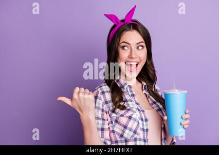 Fotoporträt einer staunenden Frau, die ein kohlensäurehaltiges Getränk in einer Hand hält und mit dem Daumen nach hinten auf einen leeren Raum blickt, der auf einer lebhaften violetten Farbe isoliert ist Stockfoto