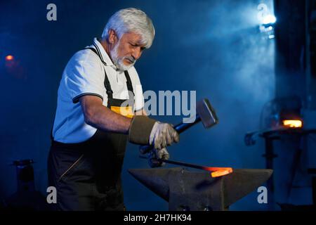 Professioneller reifer Schmied mit schwerem Hammer für die Arbeit mit geschmolzenem Stahl. Kaukasischer grauhaariger Mann in Sicherheitsuniform, der Eisen auf Amboss bei der Schmiede formt. Stockfoto