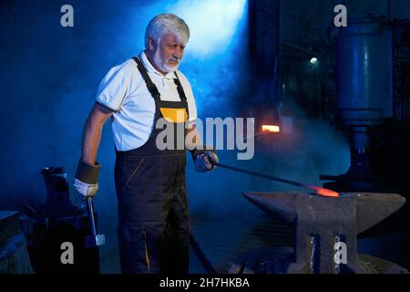 Grauhaariger kaukasischer Schmied schmiedet Stahl mit Hammer und Zepel bei der Schmiede. Reifer Handwerker mit Schürze und Handschuhen, die am Arbeitsplatz Metall Formen. Stockfoto