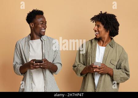 Zwei fröhliche schwarze Jungs halten Smartphones und lachen, spielen mobile Spiele zusammen Stockfoto