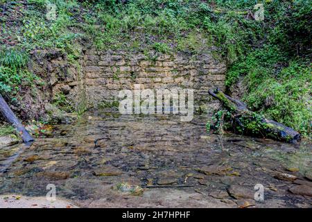 Steinmauer am Hang, umgeben von grüner wilder Vegetation, mit Ansammlung von transparentem, sauberem und kristallklarem Wasser auf Steinen auf dem g Stockfoto