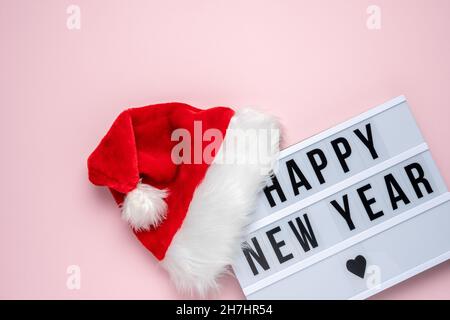 Weißer Leuchtkasten mit Text FROHES NEUES JAHR und Weihnachtsmann Hut auf rosa Hintergrund. Minimales Weihnachts- oder Neujahrskonzept. Stockfoto