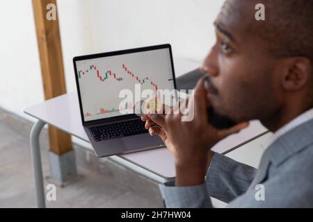 Investor mit einem traurigen und nachdenklichen Gesicht hält er eine Bitcoin und eine Ethereum-Münze in der Hand, auf dem Laptop-Bildschirm sind die Kerzendiagramme rot Stockfoto