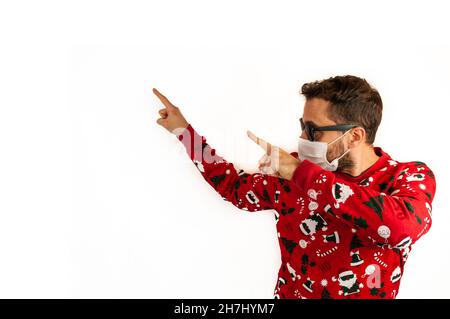 Junger weißer Mann mit rotem Weihnachtspullover und Sonnenbrille und Gesichtsmaske, isoliert auf weißem Hintergrund und Kopierraum, der mit beiden Händen auf die s zeigt Stockfoto