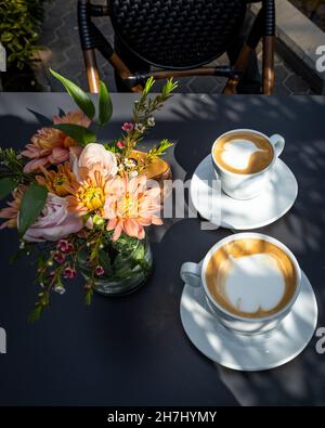 Zwei Tassen Cappuccino und eine Glasvase mit bunten Blumen auf dem Tisch im Restaurant. Leerer Stuhl, keine Leute. Stockfoto